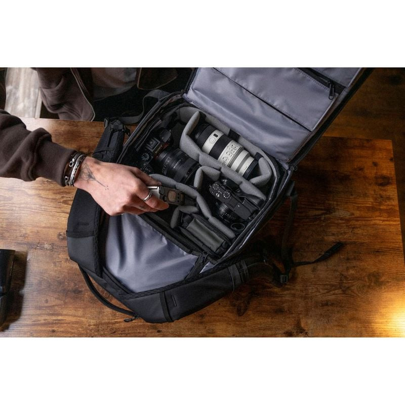 Sac à dos WANDRD PRVKE 21L ouvert sur table en bois, mains organisant appareil photo, objectifs et a