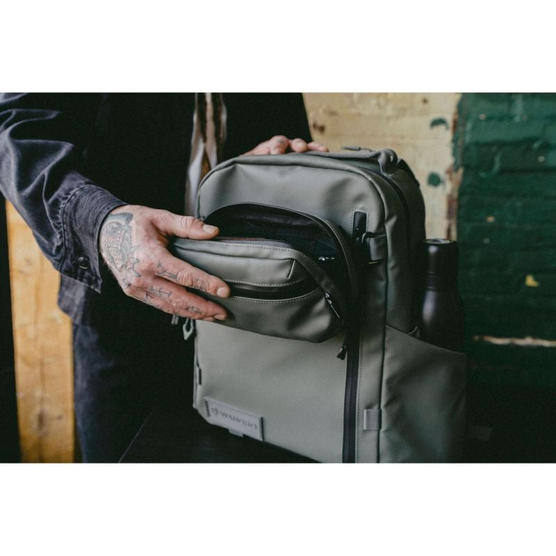Main accédant à la petite poche zippée du sac à dos WANDRD PRVKE 21 Zip vert contre un mur de brique