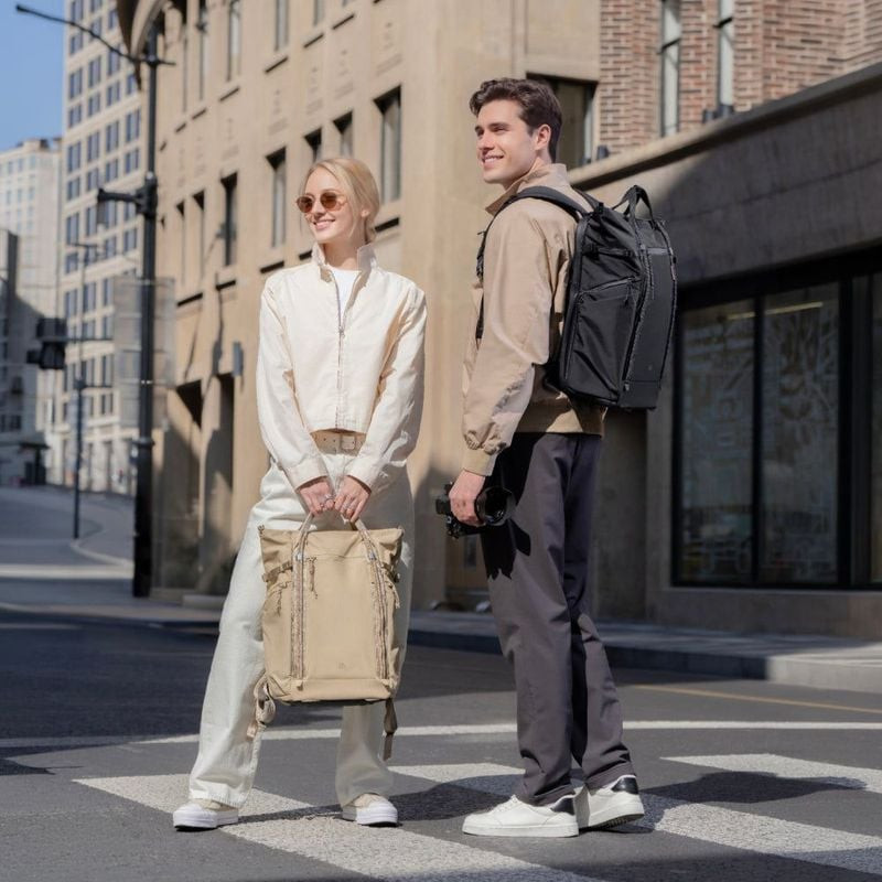 Femme portant le PGYTECH OneGo Roamer Tote 18L Sable à la main, tandis qu'un homme porte un sac à do